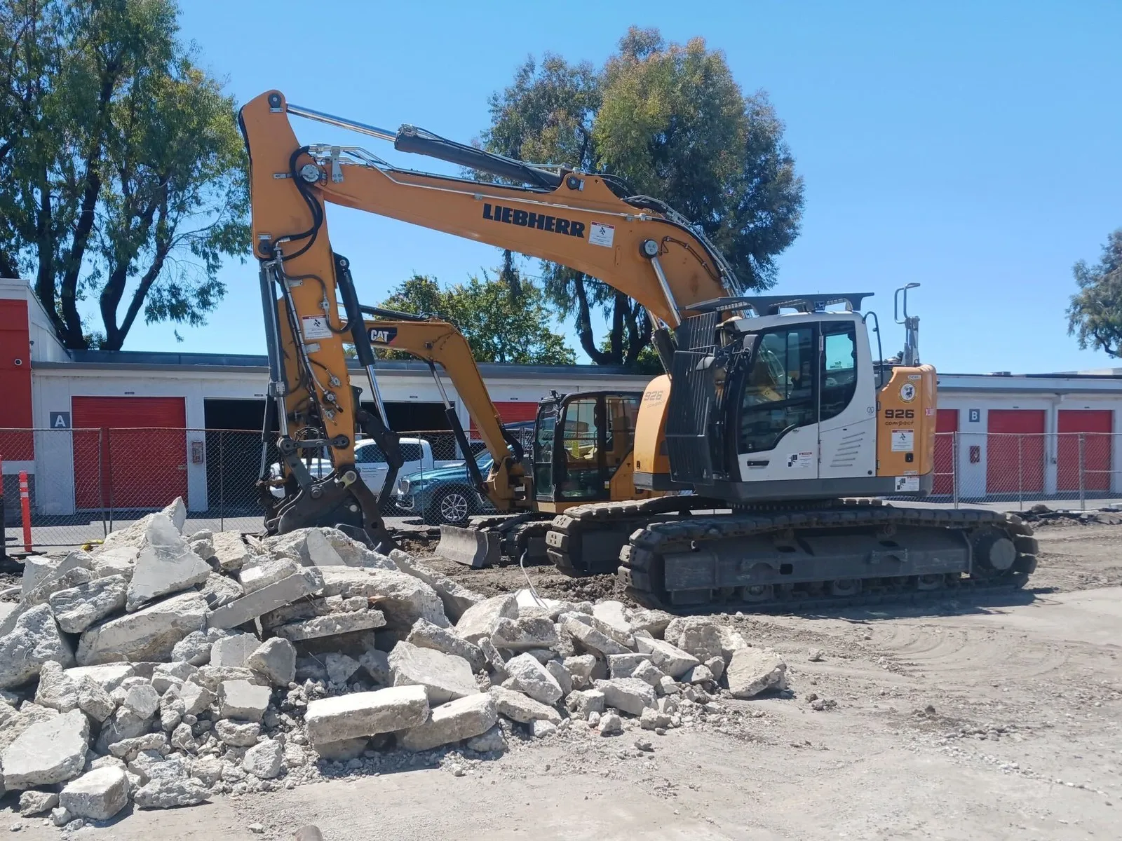 A pile of broken concrete slab in front of two excavators on a commercial demolition site, with a row of storage units behind a chain-link fence.