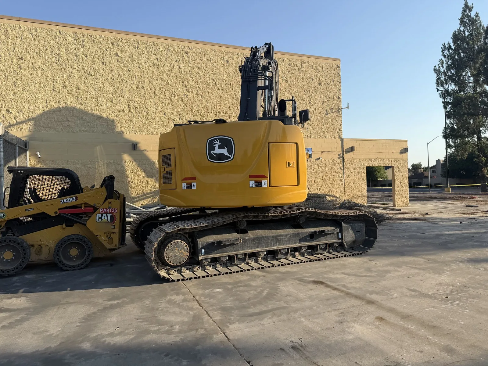 A tracked excavator and a skid steer loader parked beside a tan block wall on a demolition site, with a chain-link fence and caution tape in the background.
